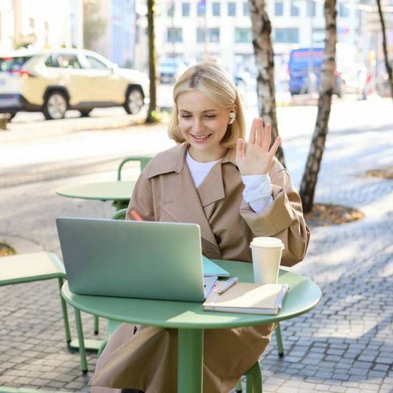 Cheerful girl with laptop, saying hello, connects to online meeting, waving hand at camera, chatting with someone via internet, sitting in outdoor coffee shop.