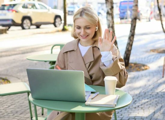 Cheerful girl with laptop, saying hello, connects to online meeting, waving hand at camera, chatting with someone via internet, sitting in outdoor coffee shop.